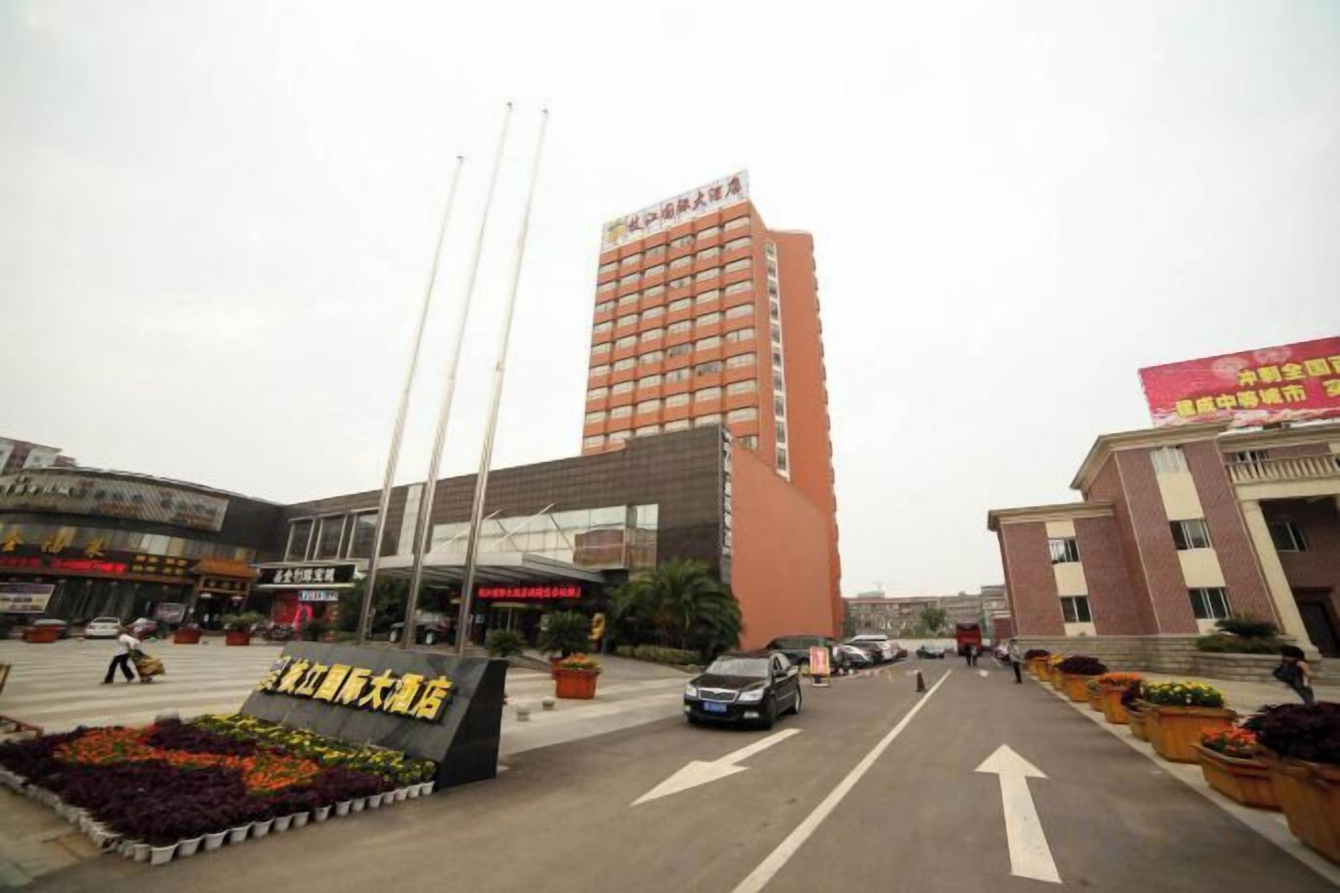 Entrada de hotel con un edificio alto al fondo, un estacionamiento y un parterre.