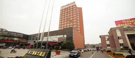 Entrada de hotel con un edificio alto al fondo, un estacionamiento y un parterre.