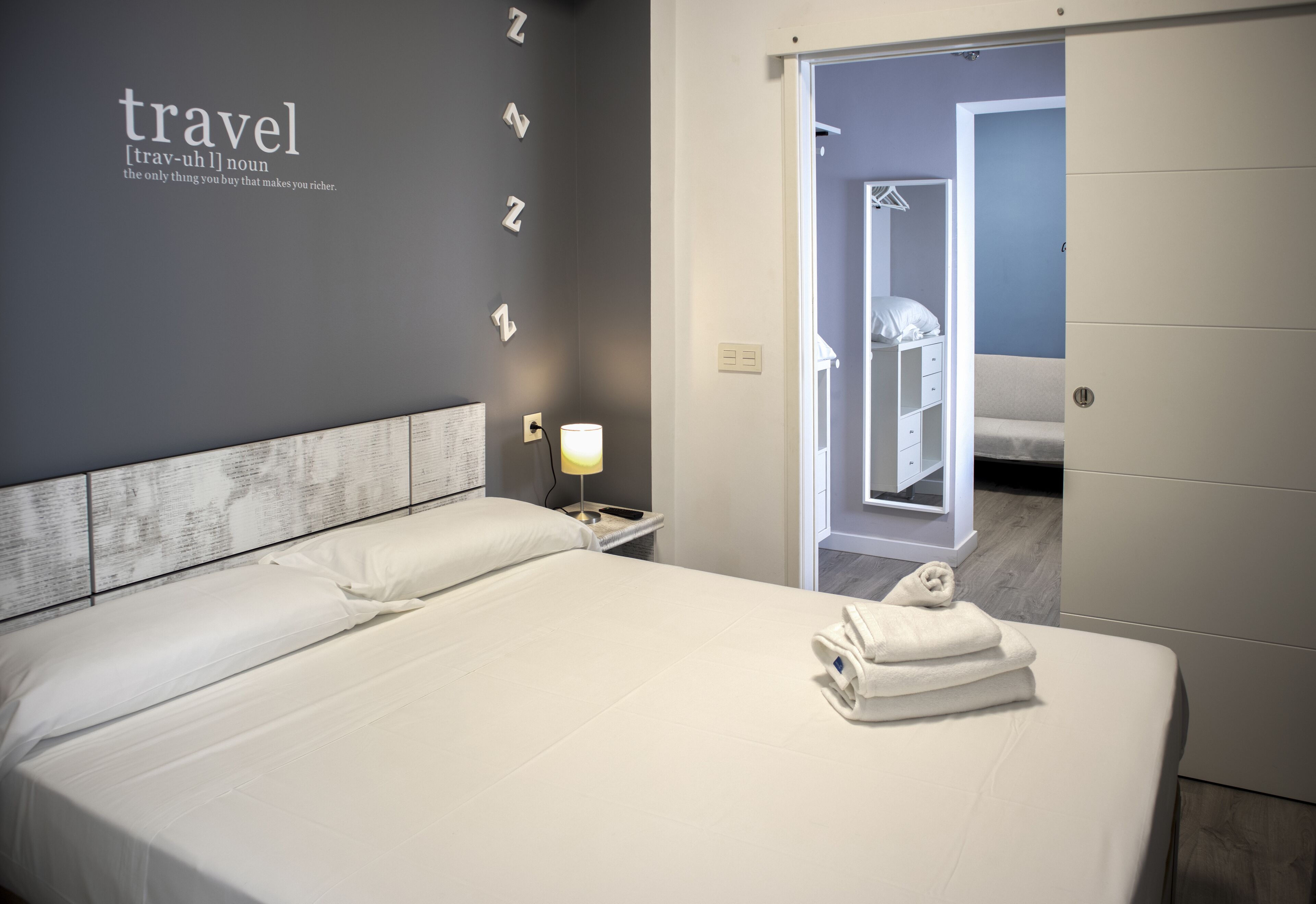 A neatly made bed with white linens and a folded towel, a bedside lamp, and a wall with the word "travel" and a definition.