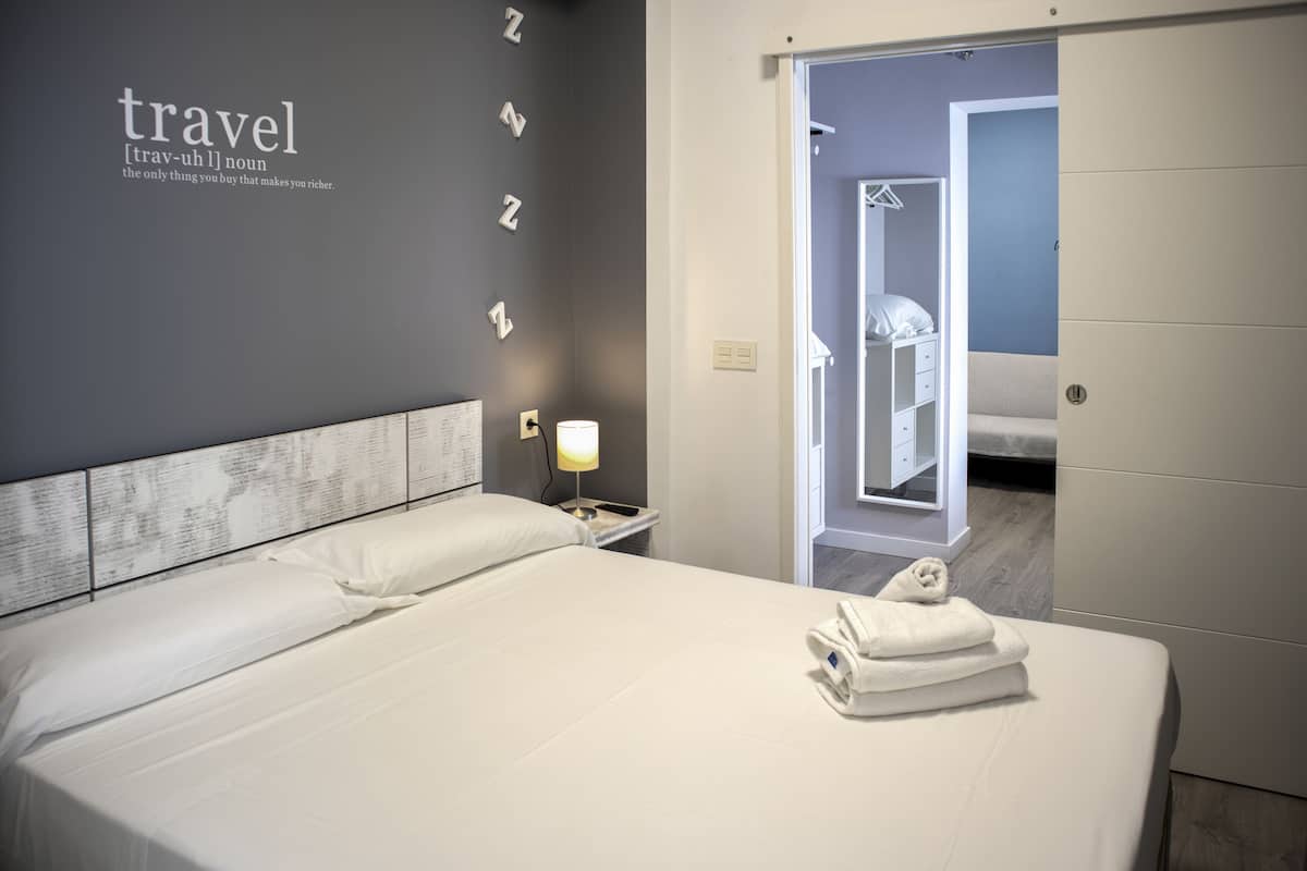 A neatly made bed with white linens and a folded towel, a bedside lamp, and a wall with the word "travel" and a definition.