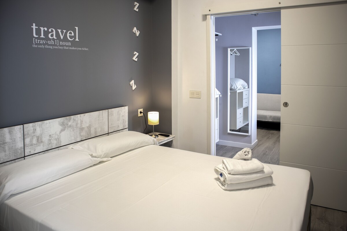 A hotel room with a large bed, a bedside lamp, a wall with the word 'travel' and a definition, a mirror, and a door leading to another room.