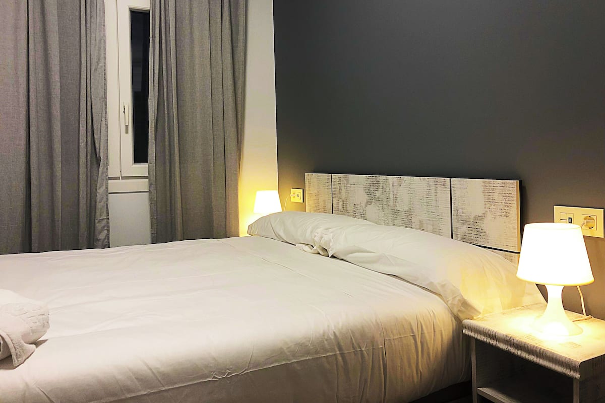 A neatly made bed with a white comforter and pillows, a bedside table with a lamp, and a window with curtains.
