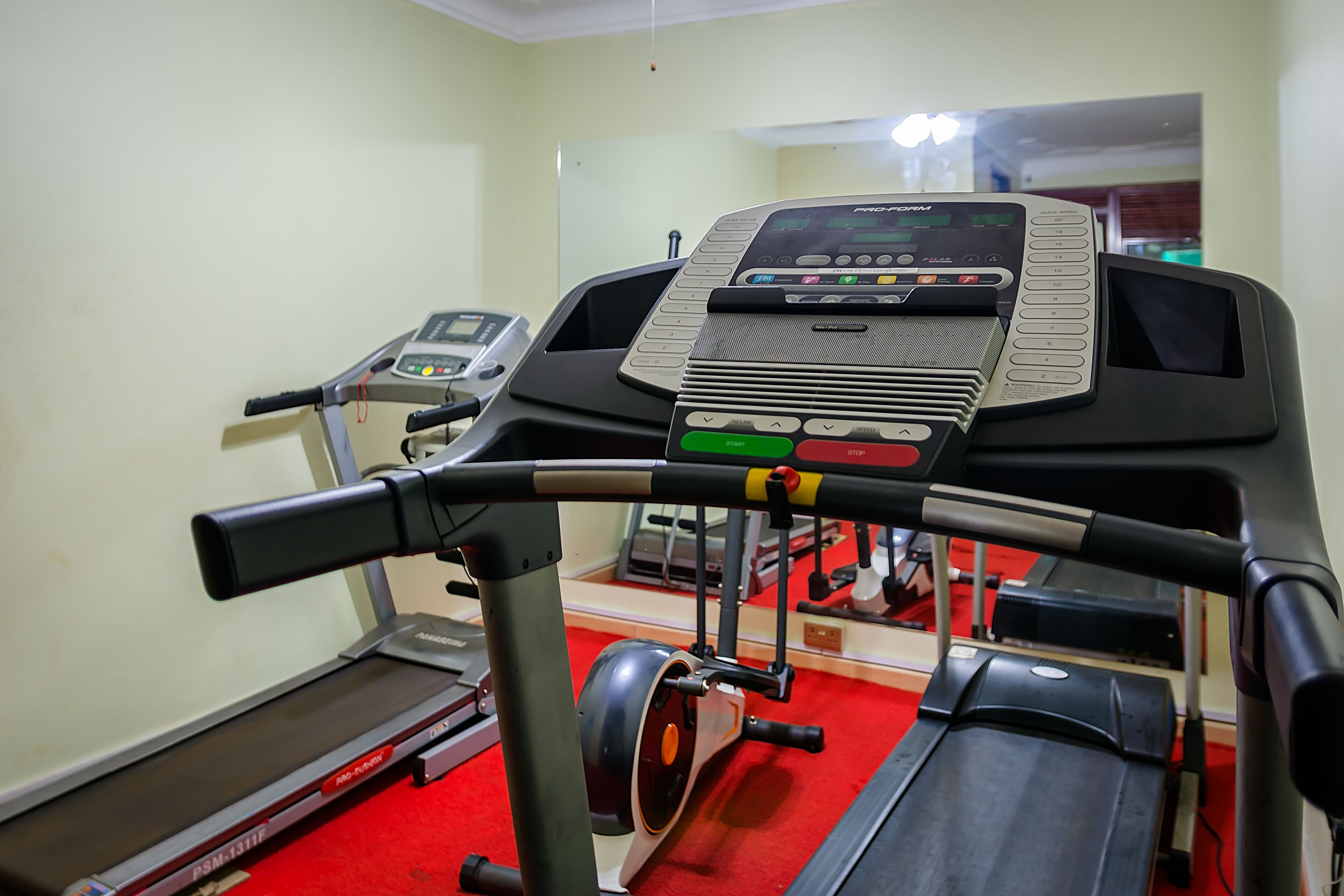 A treadmill with a digital display and control panel, a red carpeted floor, and a mirror on the wall.
