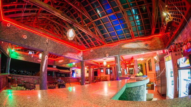 An indoor bar area with a high ceiling, wooden beams, and a glass enclosure. There are chairs and tables, and a refrigerator is visible in the background. - Chocolate Luxury Hotel (Kampala)