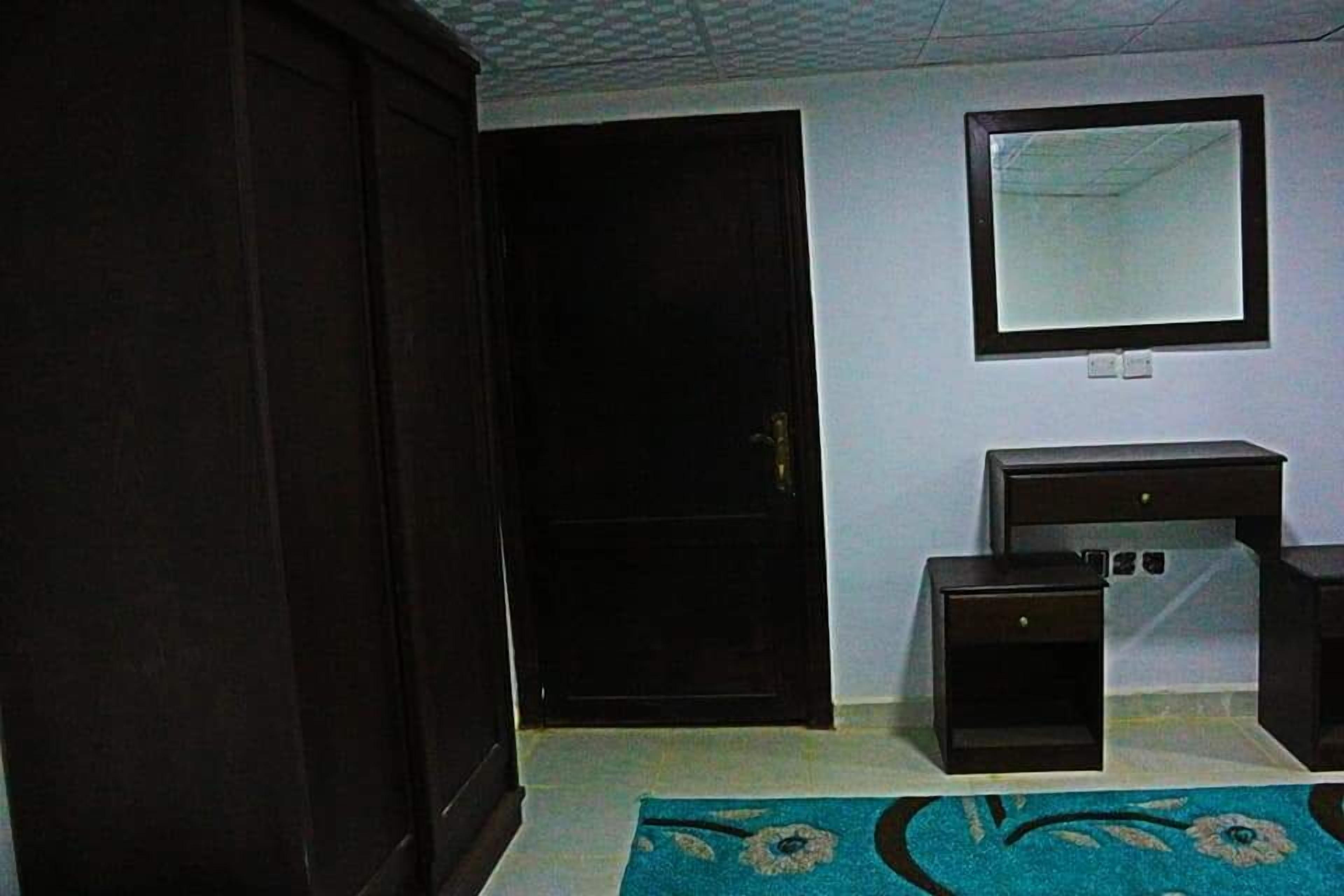 A room with a closed door, a small desk with drawers, a mirror on the wall, and a blue patterned rug on the floor.