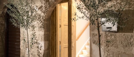Eingang eines Steingebäudes mit einer Holztür, Kübelolivenbäumen und einem Schild mit der Aufschrift 'ALLEY 7'.