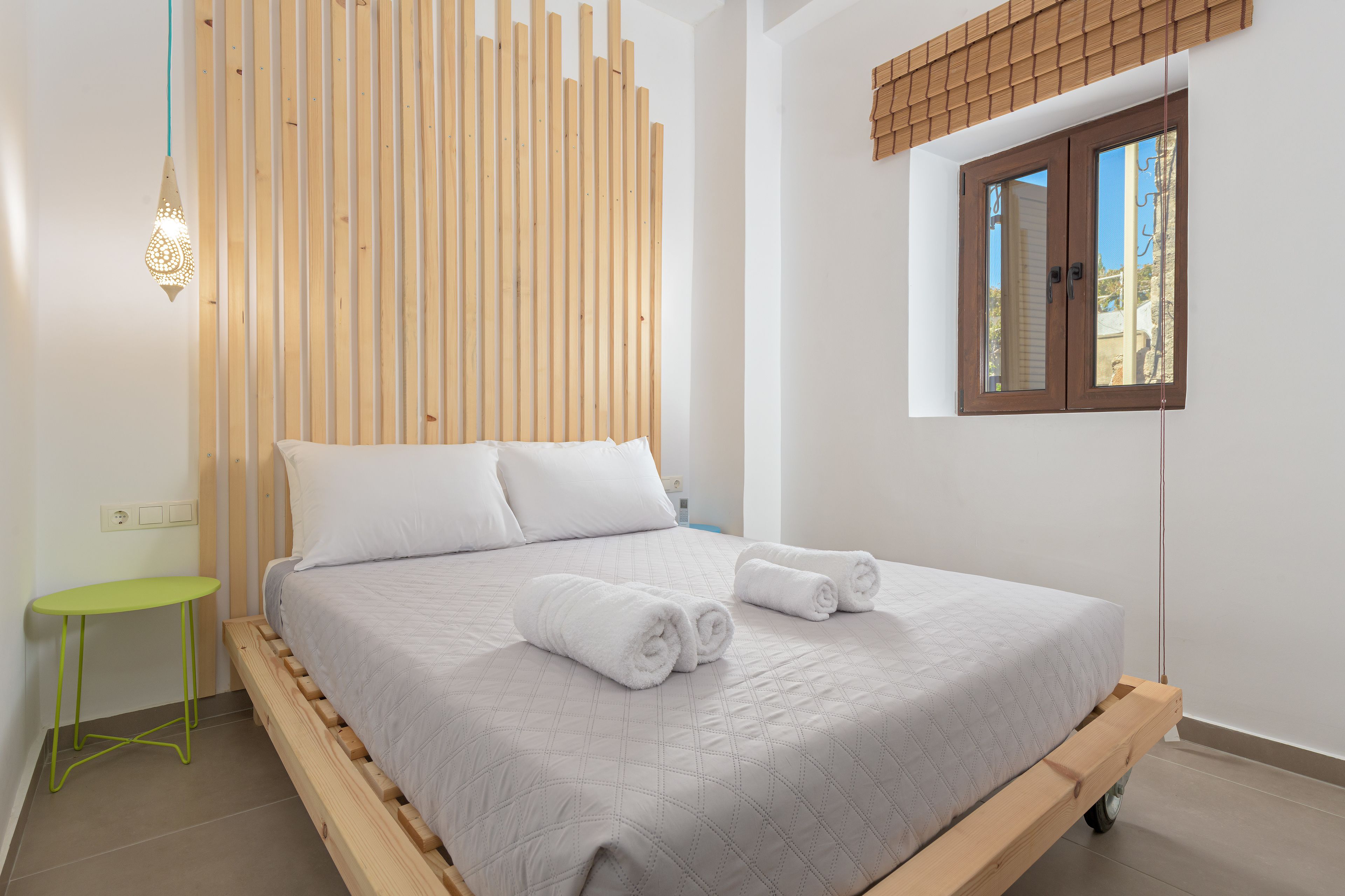 A neatly made bed with rolled towels, a small green table, and a wooden slat wall panel.