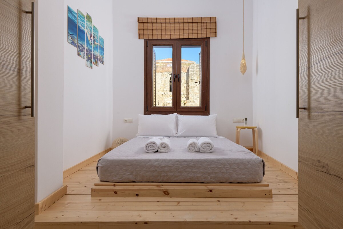 A room with a bed, a window, a small table, and a wall-mounted light fixture.