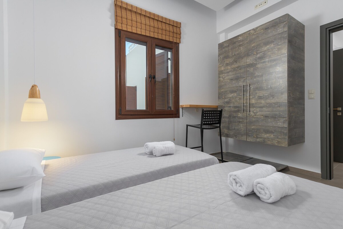 A bedroom with two beds, a wardrobe, a window with blinds, a chair, and a lamp.