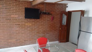 A room with a brick wall, a mounted television, a red chair, a glass table, and a refrigerator. - Casa Liceo (Guadalajara)
