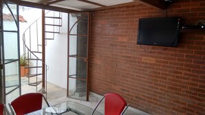 A room with a brick wall, a TV mounted on it, a glass door leading outside, and two red chairs. - Casa Liceo (Guadalajara)