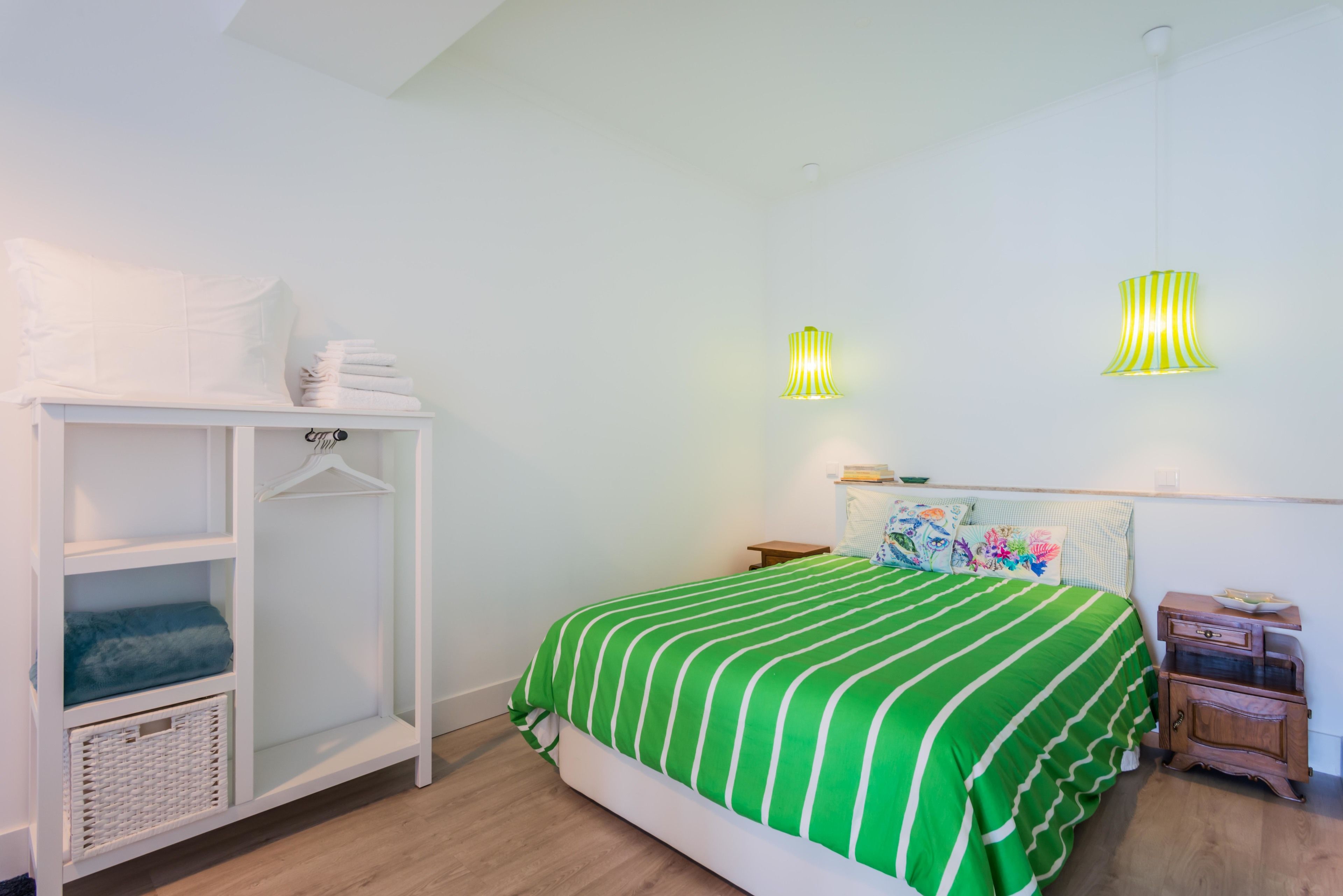 A bedroom with a green-striped bedspread, a white wardrobe, and two wall-mounted lamps.
