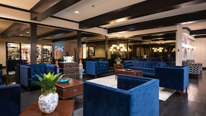 Eine Lobby mit blau gepolsterten Sitzmöglichkeiten, einem Couchtisch und einer Pflanze. Im Hintergrund ist ein Geschäft zu sehen, sowie ein Empfangsbereich mit einem Chandelier.