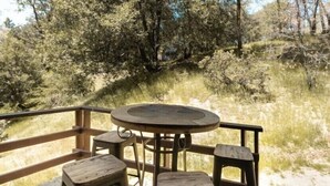 A wooden deck with a round table and four stools, surrounded by a railing, overlooking a natural landscape with trees and grass. - Cozy Treetop A-Frame, Fireplace, Arcade Games, Record Player (Lake Arrowhead)