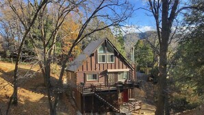 A cabin with a wooden exterior, a deck, and a staircase leading to the entrance. - Cozy Treetop A-Frame, Fireplace, Arcade Games, Record Player (Lake Arrowhead)