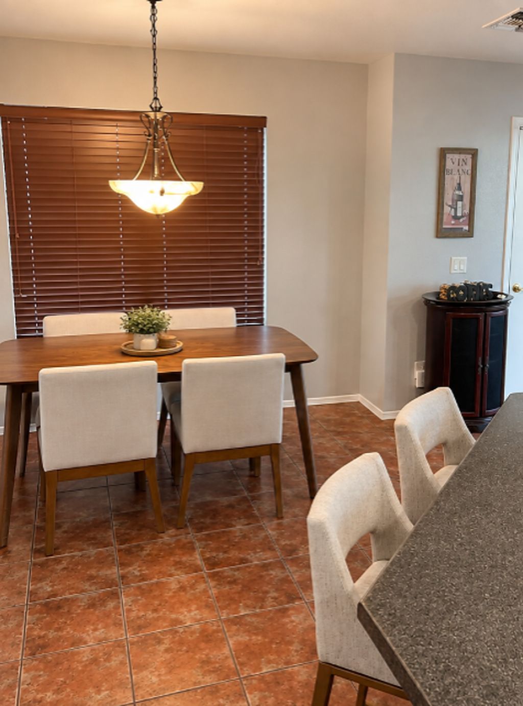 A dining area with a wooden table, four chairs, a hanging light fixture, and a potted plant.