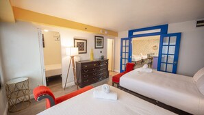 Ein Hotelzimmer mit zwei Betten, einem Schminktisch, einem roten Sessel, einem kleinen runden Tisch und einem blau gerahmten Fenster.