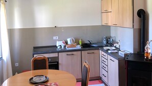 A compact kitchen with wooden cabinets, a black stove, and a dining area with a round table and chairs. - Beautiful wooden house nearby pines forest (Žabljak)