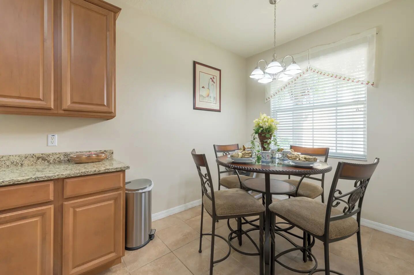 A kitchen with wooden cabinets, a granite countertop, a stainless steel trash can, a dining table with four chairs, and a window with blinds.
