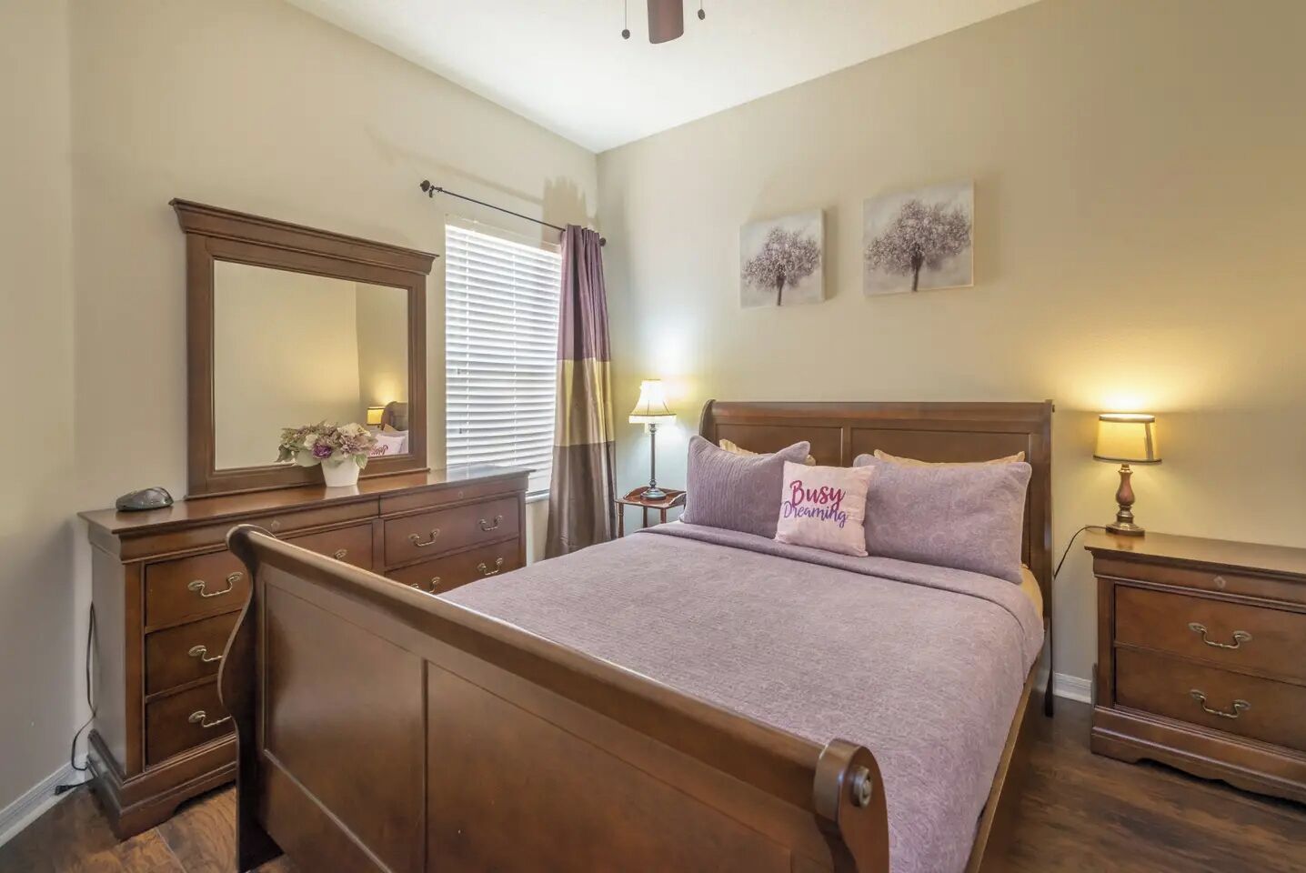 Un dormitorio con una cama de madera, una mesita de noche con lámpara, un aparador, un espejo y dos cuadros en la pared.