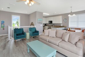 Uma sala de estar moderna com um sofá bege, poltronas verde-água e uma mesa de centro azul claro. Cozinha com armários brancos e micro-ondas visível ao fundo.