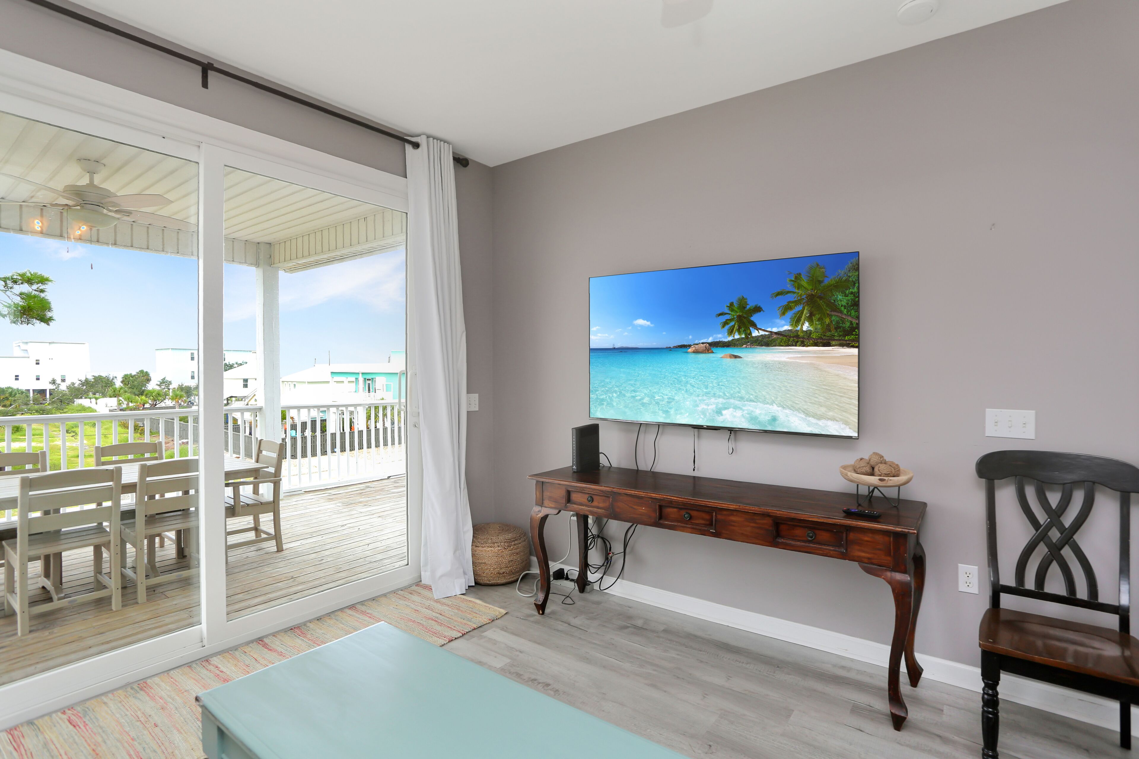 Une pièce avec un grand téléviseur fixé au mur, un bureau en bois, une chaise et une vue sur un balcon aménagé avec du mobilier d’extérieur.