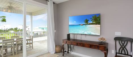 Une pièce avec un grand téléviseur fixé au mur, un bureau en bois, une chaise et une vue sur un balcon aménagé avec du mobilier d’extérieur.