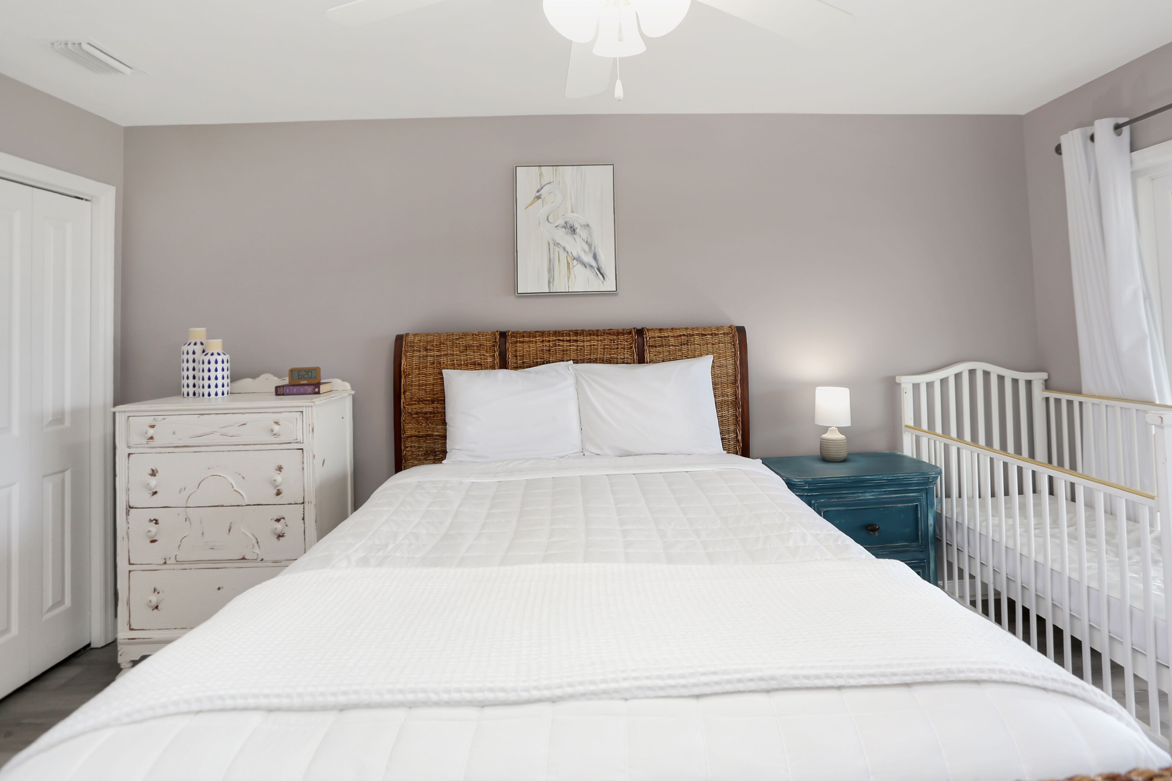 Ein Schlafzimmer mit einem Bett, einem Schminktisch, einem Nachttisch, einem Babybett und einem an der Wand hängenden Bild.