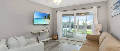 Una sala de estar moderna con ventilador de techo, televisor de pantalla plana, sofá y vista a un balcón con mobiliario exterior.