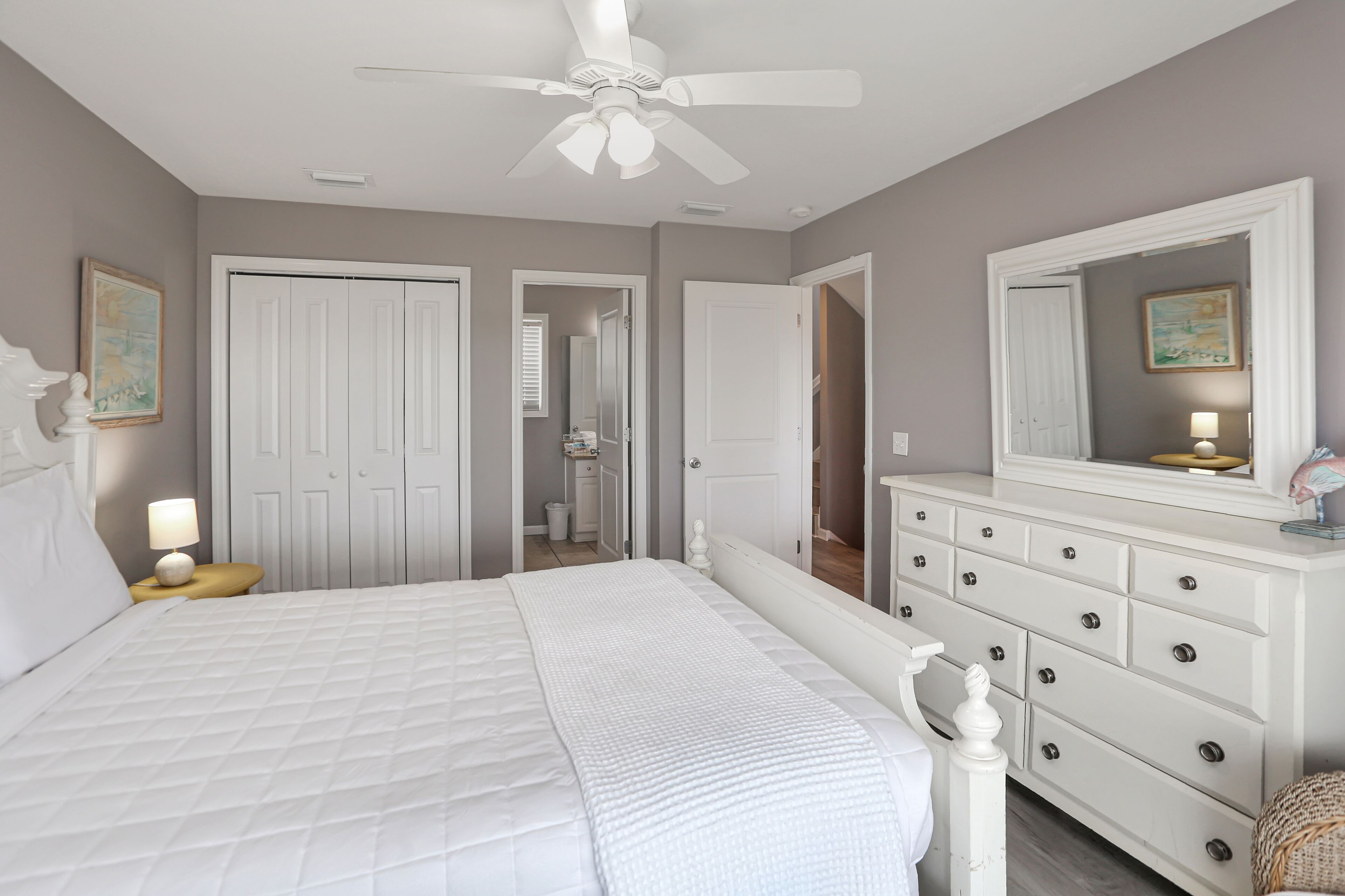 A bedroom with a large bed, a dresser, a ceiling fan, and a door leading to a bathroom.