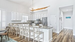 Een moderne keuken met witte kasten, een centraal eiland met barkrukken, een kroonluchter en een bordje met de tekst 'BEACH LIFE'.