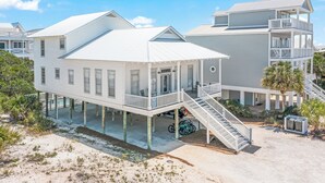 Een huis aan het strand met een balkon, een overdekte veranda en een trap naar de bovenverdieping.