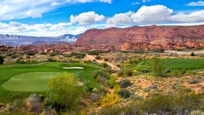 Un parcours de golf situé en plein désert, avec une chaîne de montagnes en toile de fond, un club-house et un ciel dégagé.