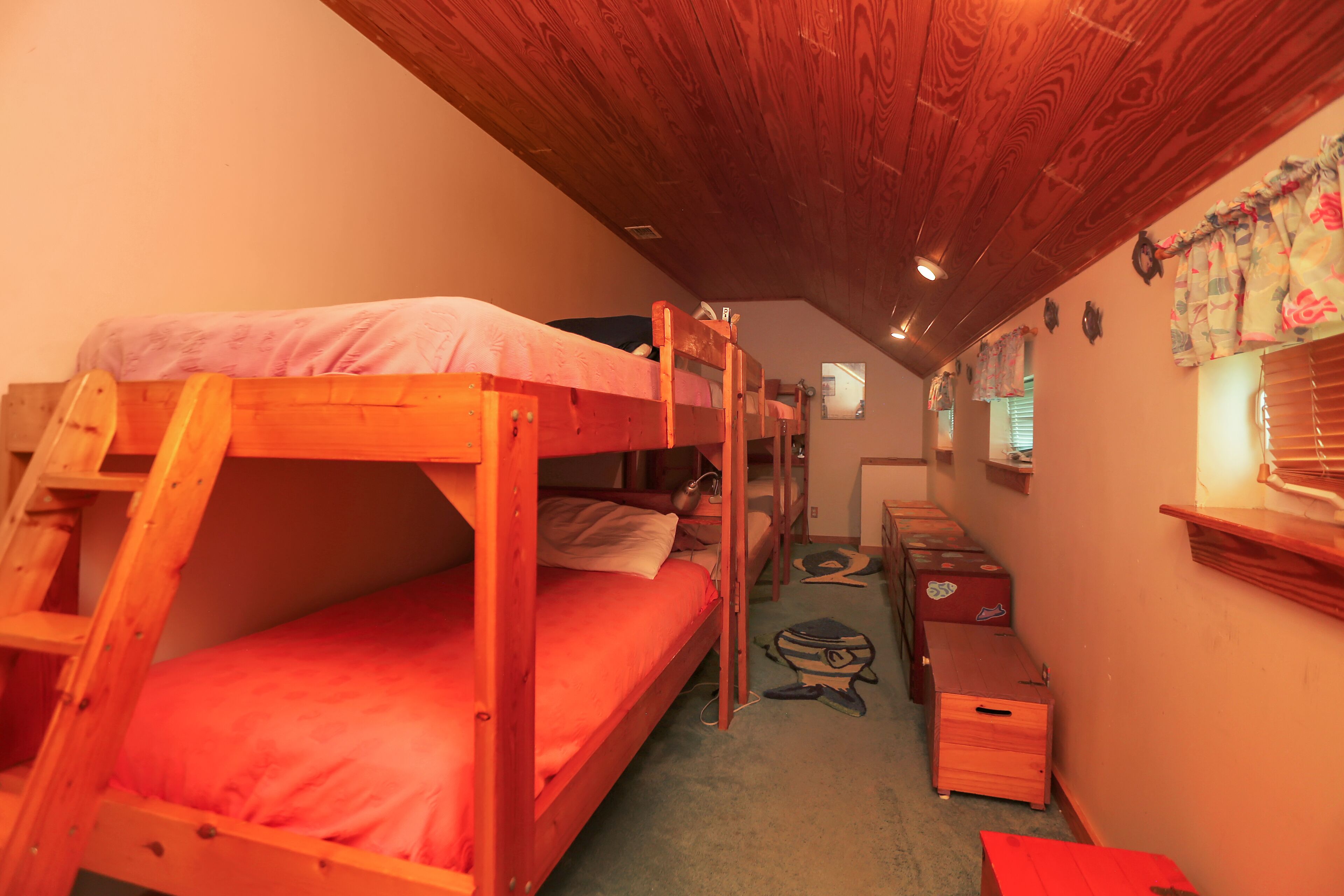 A dormitory room with bunk beds, a wooden ladder, a desk, and a small window with curtains.
