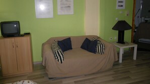 Olohuoneessa on beige sohva, televisio puukehikkoisessa kaapissa ja pieni pöytä, jossa on lamppu.