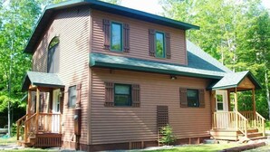 A two-story wooden cabin with a porch and a small deck. - Island Fox Nestled in the Woods on Madeline Island (La Pointe)