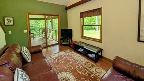 A living room with a brown leather sofa, a glass coffee table, a television, and a large window with a view of greenery. - Island Fox Nestled in the Woods on Madeline Island (La Pointe)
