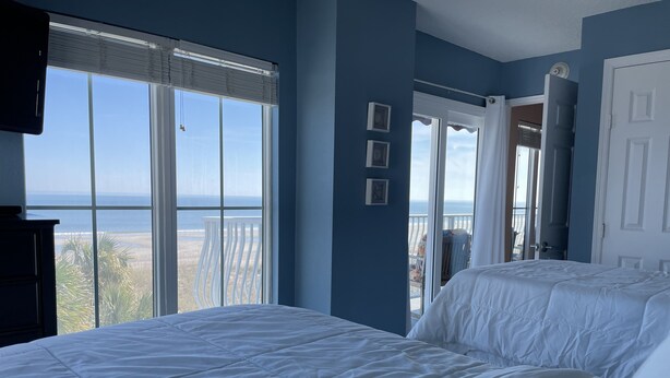 A bedroom with a large bed, a TV, and a view of the beach and ocean through the window. - Beachfront 2-bedroom condo with breathtaking ocean views from every room! (Fernandina Beach)