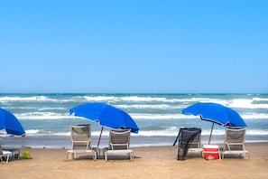 Ein Strand mit vier Metall-Strandstühlen und blauen Sonnenschirmen, wobei einer mit einem schwarzen Tuch bedeckt ist.
