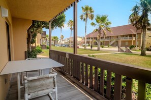 A balcony with a table and chair, overlooking a street with palm trees and a building with balconies.