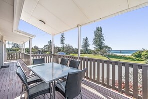An outdoor deck with a glass table and chairs, overlooking a beach and greenery.