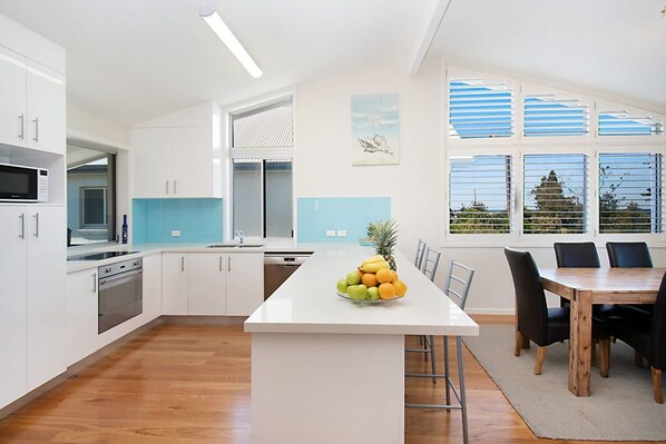 A modern kitchen with white cabinets, a central island with fruit, and a dining area with a wooden table and chairs.