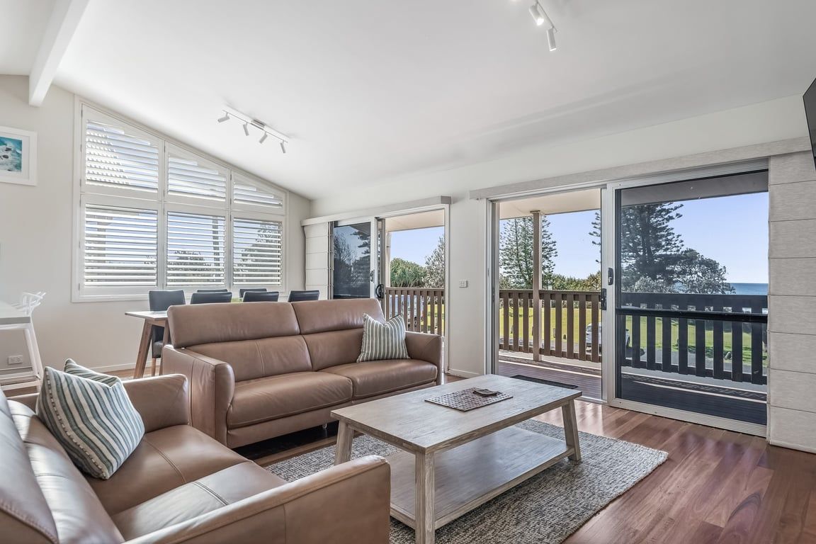 Un vaste espace de vie comprenant un canapé en cuir marron, une table basse en bois et une table à manger avec des chaises. De grandes fenêtres offrent une vue sur l’extérieur.