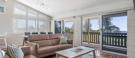 A spacious living area with a brown leather sofa, a wooden coffee table, and a dining table with chairs. Large windows provide a view of the outdoors.