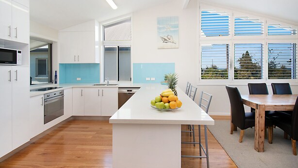 Een moderne keuken met witte kasten, een centraal eiland met fruit, een eethoek met een houten tafel en stoelen, en grote ramen met zonwering.