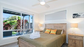 Een slaapkamer met een groot raam, een bed met houten hoofdeinde, twee nachtkastjes met lampen en een klein houten kastje.