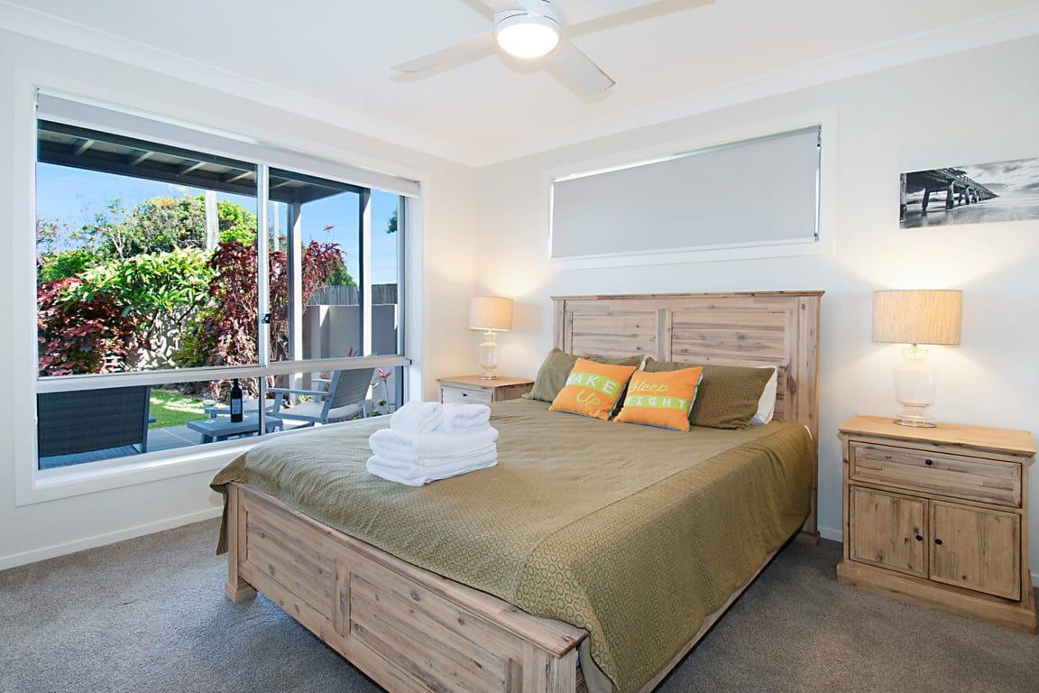 A bedroom with a large window, a bed with a green bedspread, two bedside lamps, and a wooden headboard.