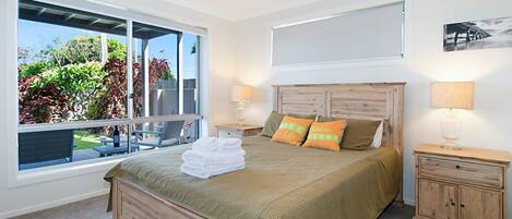 A bedroom with a large window, a bed with a green bedspread, two bedside lamps, and a wooden headboard.