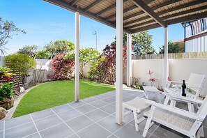A covered patio area with a tiled floor, white lounge chairs, and a small table with a bottle and glass.