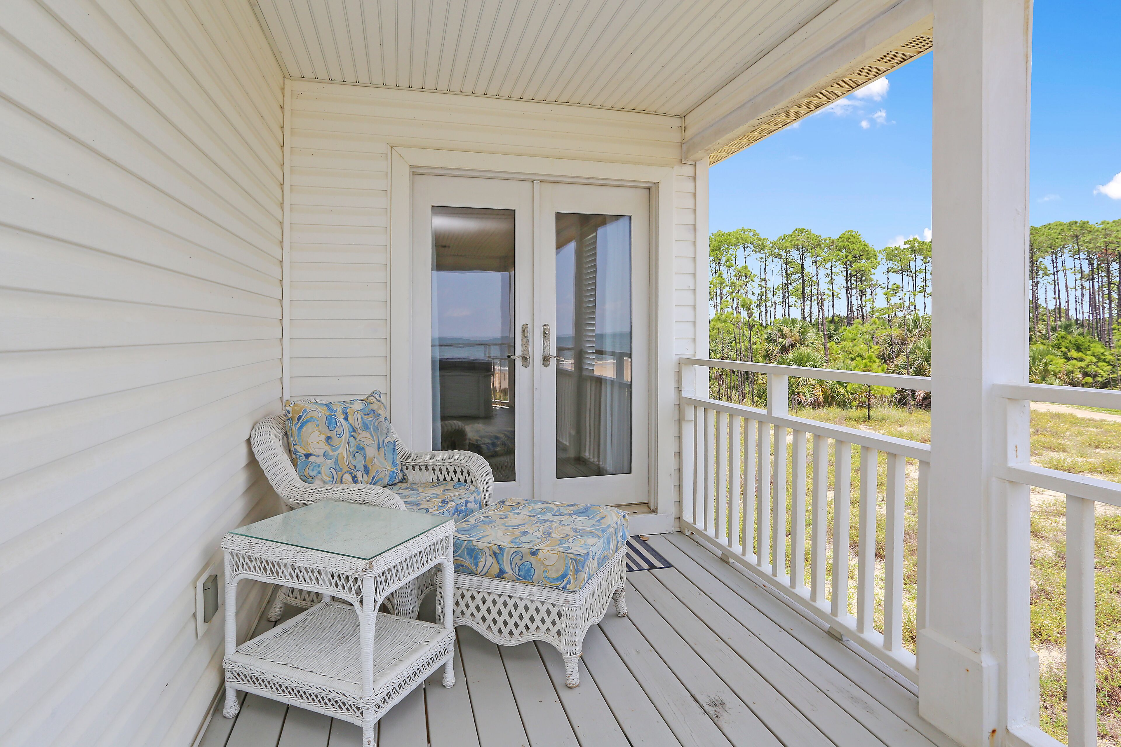 A covered porch with white railings, a glass door, wicker furniture, and a view of a forest.